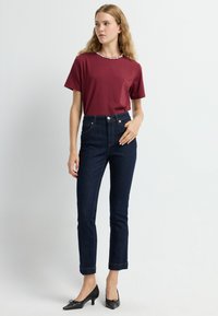 Bordová trička s krátkým rukávem a dekorativním výstřihem, ve spojení s tmavě modrými džínami s vysokým pasem a černými podpatky, předváděné modelem.