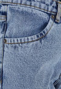 Pantalones vaqueros de mezclilla azul claro con un diseño clásico de cinco bolsillos, con costuras amarillas y una superficie texturizada que muestra un ligero desgastado.