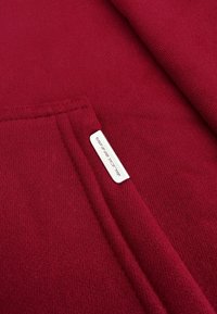 Rode sweatshirt met een gestructureerde stof, een kangoeroezak en een klein wit label erin genaaid, dat het merk aangeeft.