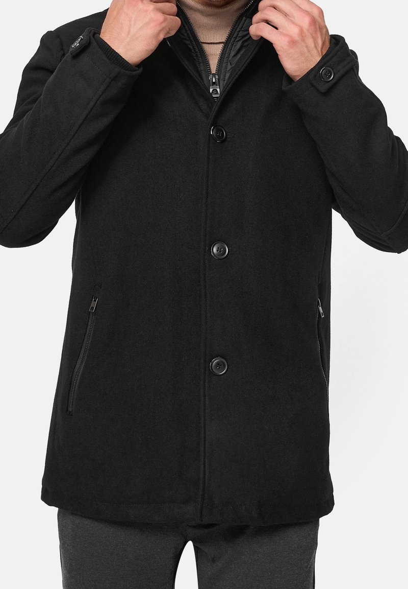 Homme portant un manteau noir boutonné avec des poches zippées et un col roulé beige, ajustant le col avec les deux mains.