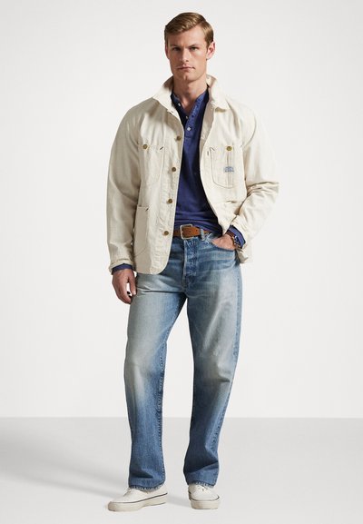 Λευκή καμβάς ζακέτα με δύο τσέπες στήθους, ναυτική μπλούζα πόλο, ανοιχτά μπλε τζιν και μπεζ παντόφλες. Μινιμαλιστικός σχεδιασμός, casual εφαρμογή.