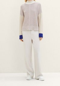 Frau trägt einen beigen und cremefarbenen Strickpullover mit blauen Bündchen, lockere weiße Hosen und weiße Sneakers vor einem schlichten hellen Hintergrund.