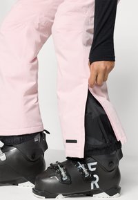 Rosa Skihosen mit einem eleganten Design und schwarzem Innenfutter, ausgestattet mit einem Reißverschluss am Saum und schwarzen Skistiefeln mit silberner Hardware.