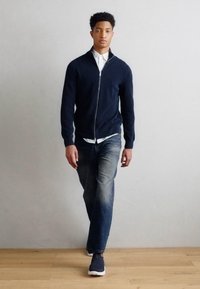 Jūras spēku zip-up kardigāns, balts apkakles krekls un izbalējuši zili džinsi. Modelis nēsā jūras spēku krāsas sporta apavus. Fons ir neitrāls ar koka grīdām.