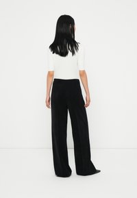 Schwarzweite Hose aus weichem Stoff mit hoher Taille und glatter Textur. Das Modell trägt ein enges weißes Oberteil, von hinten gesehen.