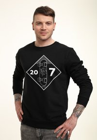 Felpa nera con una grafica a forma di diamante che presenta un design di skateboard, i numeri "20" e "7" in bianco, e maniche e orlo a costine.