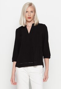 Czarna, strukturalna bluzka z dekoltem w serek, rękawami o długości trzy czwarte oraz luźnym krojem, z poziomymi fałdami i dolną krawędzią z subtelnymi wycięciami.