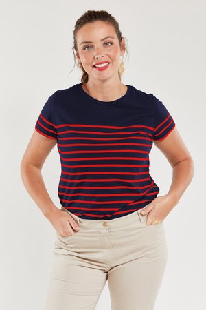 Femme souriante aux cheveux bruns attachés, portant une chemise bleu marine à rayures rouges et un pantalon beige, les mains dans les poches, fond uni.