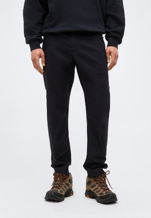 Personne portant un pantalon noir et des chaussures de randonnée marron, debout devant un fond clair uni.