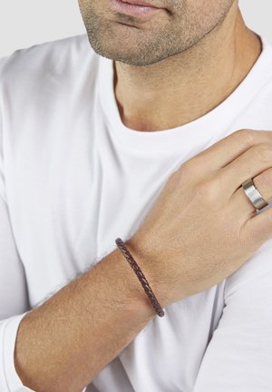 Mann trägt ein weißes Langarmshirt, ein geflochtenes braunes Lederarmband am linken Handgelenk und einen silbernen Ring an der rechten Hand.