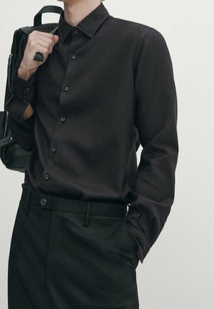 Personne portant une chemise noire boutonnée et un pantalon noir, tenant une bandoulière de sac sur une épaule avec une main dans une poche, sur un fond uni.