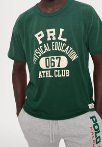 Zelené bavlněné tričko s krátkým rukávem a bílým potiskem: "PRL TĚLESNÁ VÝCHOVA 067 ATLETICKÝ KLUB." Má kulatý výstřih a volný střih.