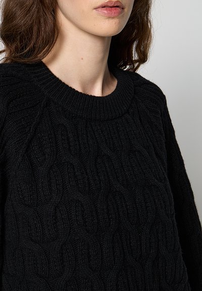 Femme à la peau claire et aux cheveux bruns ondulés, portant un pull noir en maille torsadée avec un col rond côtelé.