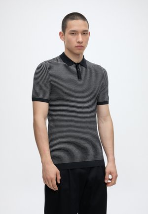 Uomo che indossa una polo a maniche corte a righe bianche e nere con colletto e polsini neri, in piedi davanti a uno sfondo chiaro e uniforme.