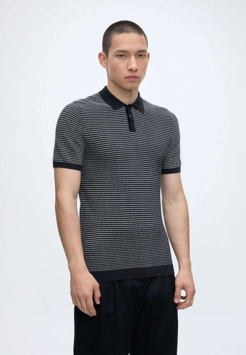 Homme portant un polo à manches courtes rayé noir et blanc avec un col et des poignets noirs, debout devant un fond uni clair.