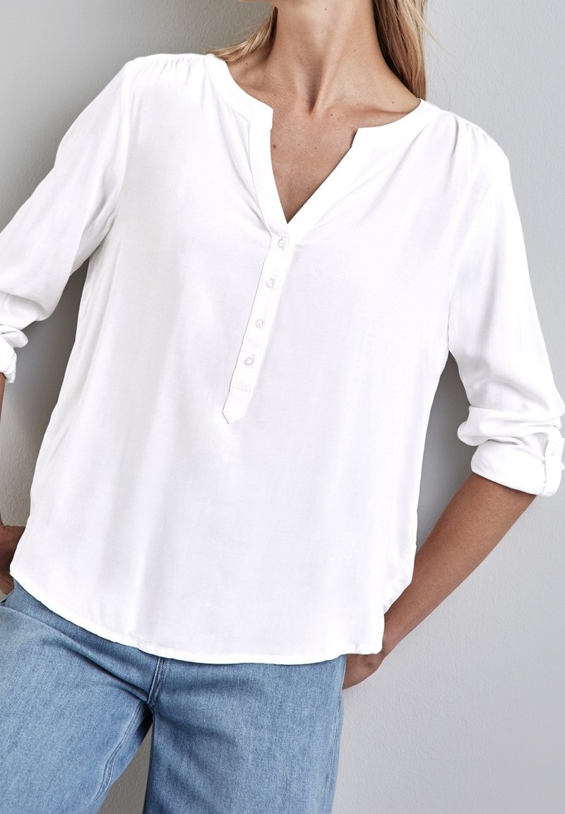 Blouse blanche à manches longues de style henley, dotée d'une patte de boutonnage avec fermeture par boutons, en tissu doux, avec poignets retroussés. Portée avec un denim clair.