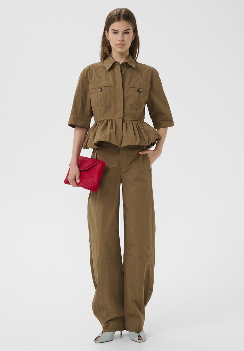 Vrouw draagt bruine wijde broekspijpen en een top met gerimpelde mouwen, houdt een rode clutch vast en draagt lichtblauwe open schoenen tegen een effen achtergrond.