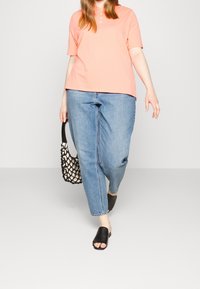 Persikofärgad polo t-shirt med knappar, avslappnade blå jeans, svarta sandals i slingback-modell och en svart vävd handväska, allt mot en neutral bakgrund.