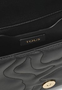 Bolso de mano de cuero negro con patrones en relieve de olas, herrajes en tono dorado y una etiqueta de marca con "TOUS" dentro del compartimento.