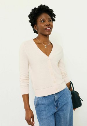 Frau mit kurzen, lockigen Haaren, die eine hellrosa, geknöpfte Strickjacke, blaue Jeans, eine silberne Halskette trägt und eine kleine schwarze Tasche hält.