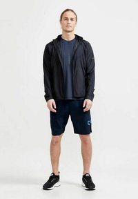 Schwarze leichte Jacke, dunkelblaues kurzärmliges Hemd, marineblaue Shorts mit Logo und schwarze Sportschuhe. Entwickelt für aktive Freizeitkleidung.