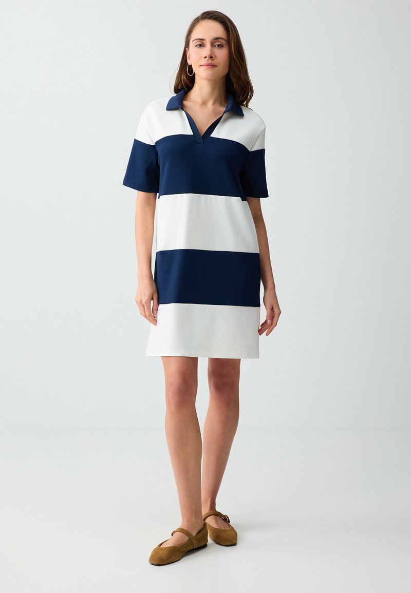 Femme debout portant une robe à manches courtes larges rayures bleu marine et blanc avec un col et des chaussures plates marron sur fond blanc.