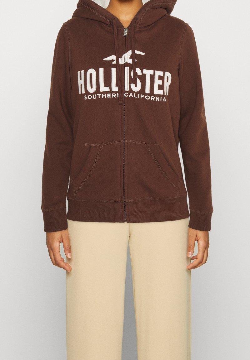 Person som bär en brun zip-up hoodie med texten "Hollister Southern California" och beige byxor, stående mot en enkel bakgrund.