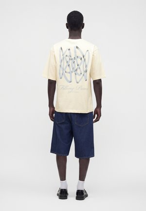 Mand iført en cremefarvet t-shirt med sølv abstrakt design og tekst på ryggen, mørkeblå knælange shorts, hvide sokker og sorte sko.