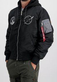 Musta bomber-takki, jossa on tilateemaisia kirjailtuja laattoja, hihassa vetoketjullinen tasku sekä resorit hihansuissa ja helmassa. Takissa on hupullinen malli.