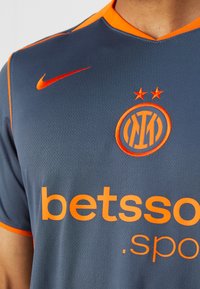 Maillot de football gris foncé avec des accents orange, présentant un col en V, le logo Nike et un emblème de club brodé avec deux étoiles au-dessus.