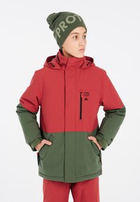 Rote und grüne wasserfeste Jacke mit Kapuze, Reißverschluss-Tasche und elastischen Bündchen. Kombiniert mit einer grünen Mütze mit auffälligem Text.