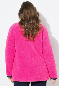 Vaaleanpunainen fleece-takki, jossa on tekstuuri ja pehmeä pinta, rento malli, pitkät hihat ja tumma reunus hihoissa. Takakatsaus, ei näkyvää metalliosaa.