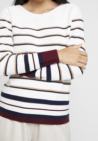 Sweter w paski w kolorze białym, z wąskimi liniami w beżowym, granatowym i bordowym. Prążkowane mankiety, okrągły dekolt i miękka faktura.