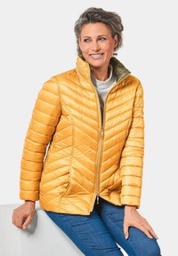 Veste en duvet jaune avec un motif en chevrons, dotée d'une fermeture à glissière sur le devant et d'un col montant. Texture lisse et poignets élastiques, associée à un jean bleu.