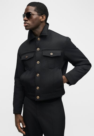 Άνδρας που φοράει μαύρο σακάκι με χρυσά κουμπιά και τσέπες με καπάκια, μαύρο παντελόνι και σκούρα γυαλιά ηλίου, κοιτώντας προς τα αριστερά.