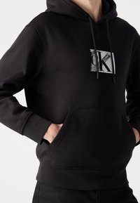 Persona con una sudadera negra con bolsillo frontal y un logo 'CK' desgastado en el pecho, con las manos metidas en el bolsillo.