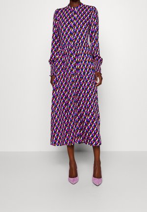 Vrouw draagt een midi-jurk met lange mouwen en knopen, met een meerkleurig geometrisch patroon, gecombineerd met lavendelkleurige pumps met puntige neus.