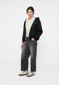 Sweat à capuche noir avec une doublure en polaire, un pull en tricot beige en dessous, un jean gris ample et des baskets beiges avec des accents marron.