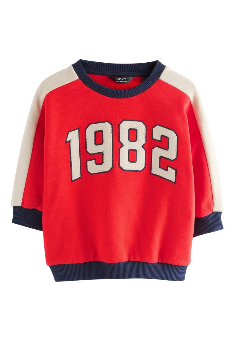 Sweatshirt rouge arborant un grand graphisme "1982" en crème et marine, avec des panneaux d'épaule crème et des poignets et ourlet côtelés marine. Texture douce.