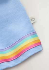 Vaate vaaleansinisestä puuvillasta, jonka helmassa on raidat purppuran, vihreän, keltaisen, oranssin ja vaaleanpunaisen sävyissä. Tunnuksessa lukee "Pieni Lintu."