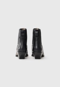 Botas de tobillo de cuero negro con un tacón afilado, que presentan una textura suave, costuras verticales y una pequeña correa en el talón como detalle.