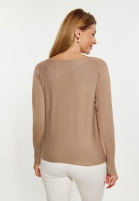 Pulover bej cu mâneci lungi, cu o croială lejeră, mansete ondulate și o decolteu larg, asortat cu pantaloni albi. Textură netedă, design minimalist.