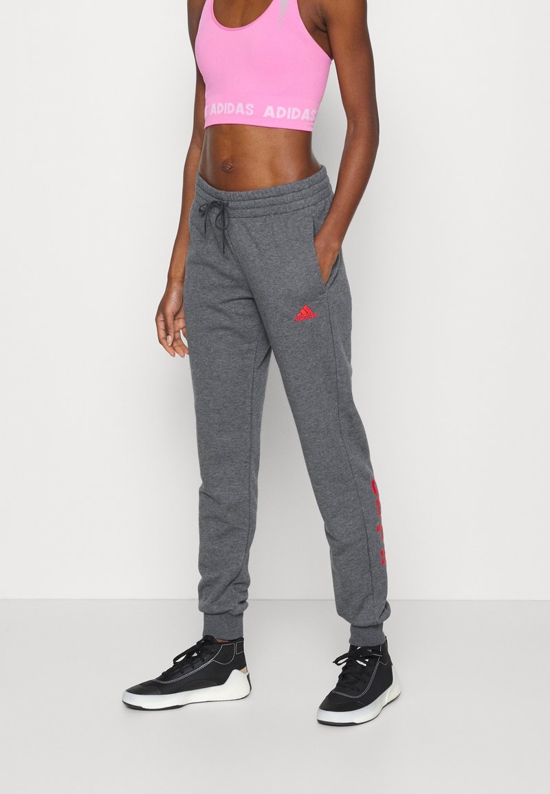 Grå joggingbukser med rødt Adidas-logo, elastisk taljebånd og manchetter, kombineret med en lyserød kort sports-bh. Sorte træningssko fuldender looket.