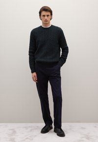 Dunkelgrüner gestrickter Pullover mit strukturiertem Zopfmuster, kombiniert mit marineblauen Hosen und schwarzen Schuhen. Das Model steht vor einem neutralen Hintergrund.