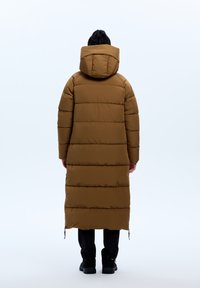 Bruine gewatteerde lange jas met een capuchon, voorzien van horizontale gestikte secties en ritssluitingen onderaan. Draagt over zwarte broek en schoenen.