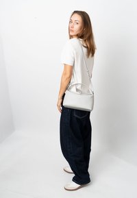 Grey leather handbag with a curved handle and adjustable strap. Model wears loose blue jeans and white sneakers, set against a plain backdrop.