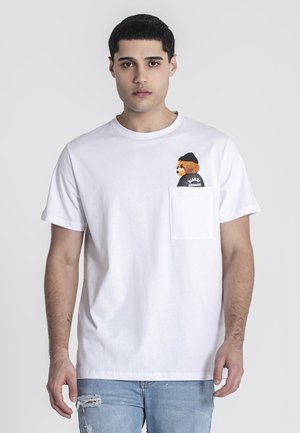 T-shirt bianca in cotone con maniche corte, dotata di una tasca sul lato sinistro del petto e un grafico di un orso che indossa un berretto. Jeans blu forati.