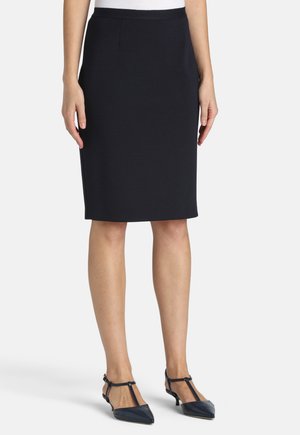 Ženska v črni svinčniki krilu dolžine do kolen in črnih čevljih z nizko peto in T-trakom, stoji pred belim ozadjem.