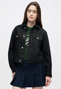 Melnas džinsa jaka ar sudraba pogām, krūšu kabatām un dzeltens diegu šuvēm, kārtaina pār zaļu adītu džemperi un tumši zilu kroku svārku.