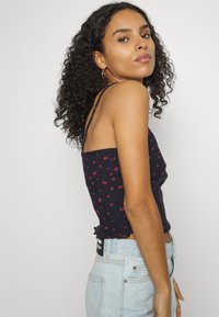 Top cortado navy com alças finas, apresentando um padrão pequeno de cerejas vermelhas e uma textura canelada. Detalhe de bainha franzida. Calças de ganga de cintura alta em azul claro.